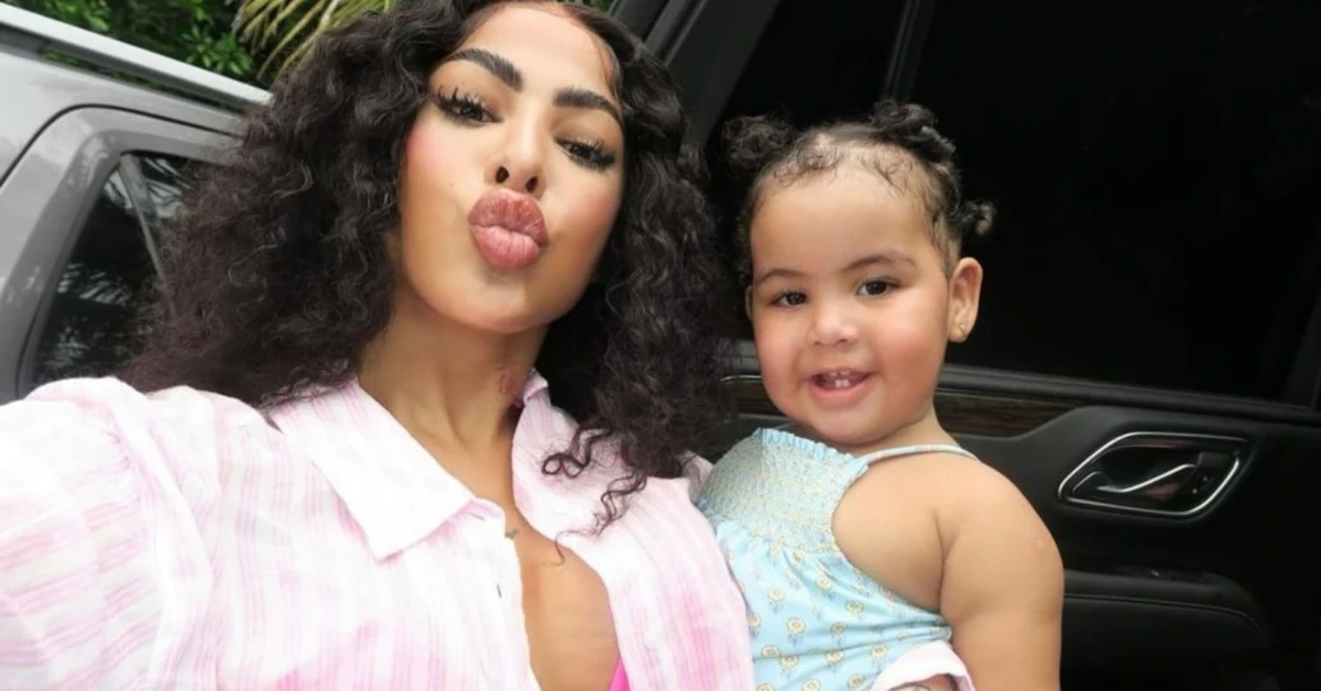 Woman with curly hair in a pink striped shirt pouts for a selfie next to a smiling toddler in a blue dress inside a car interior.