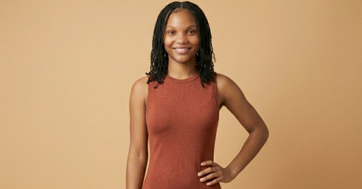 Smiling woman with braided hair stands against a beige backdrop wearing a rust-colored sleeveless top, hand on hip.