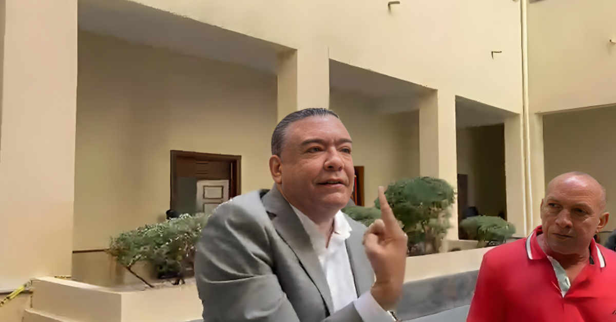 Two men stand in a pale beige courtyard; the man in a gray suit gestures with his index finger raised while speaking, as a bald man in a red polo looks on.