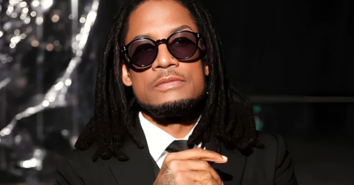 Man with dreadlocks wearing a black suit, white shirt, and tie, sporting dark sunglasses and clasping his hands in front against a dark background.