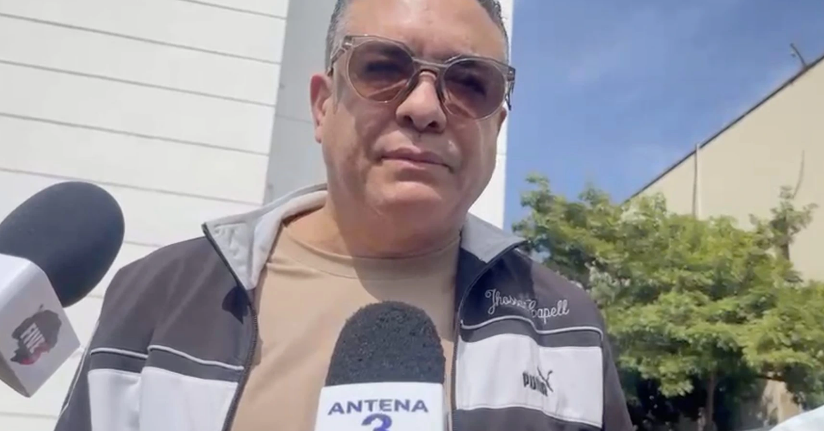 Man wearing sunglasses and a beige shirt being interviewed outdoors, holding a microphone with ANTENA 3 logo, in front of a light building and trees.