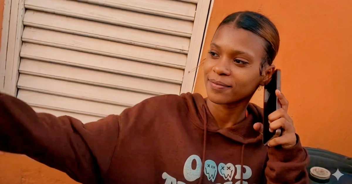 Woman in a brown hoodie talking on a smartphone outdoors by an orange wall.