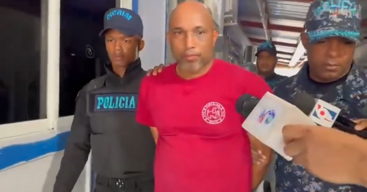 Man in a red shirt is escorted by two police officers as reporters hold microphones nearby at the scene.”, “A man in a red T-shirt being led through a hallway by police while media cameras record.”, “Two officers flank a man in a red shirt; one wears a POLICIA patch and microphones are in the frame.”, “Police escort the man in a red shirt as reporters with microphones cover the incident.”, “A man in a red shirt walks between two officers with media microphones in view.”