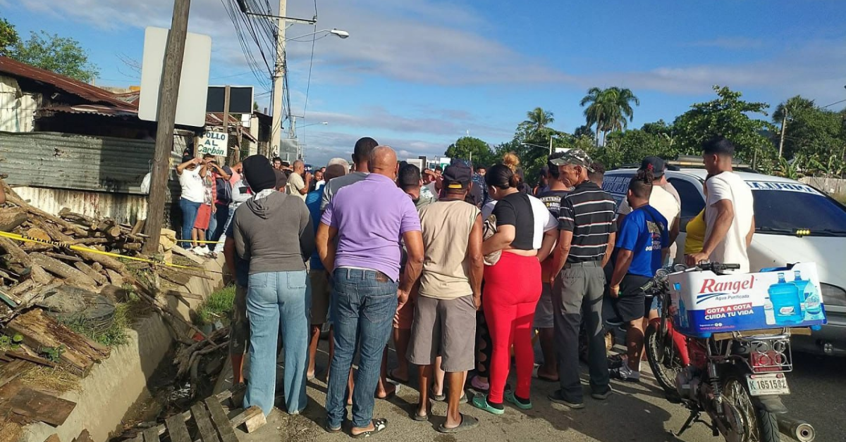 Large crowd of people gathered on a street beside a collapsed wall and debris, police tape marking off the area, under a clear blue sky.