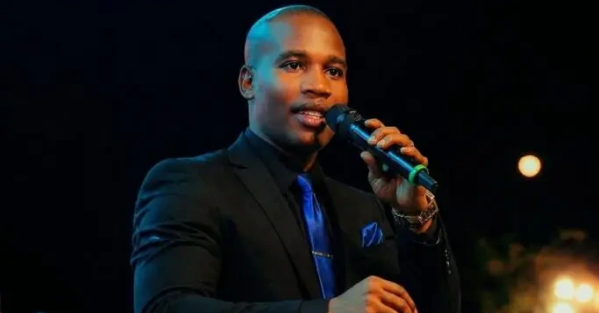 Man in a black suit and blue tie speaking into a handheld microphone on a dark stage at night.
