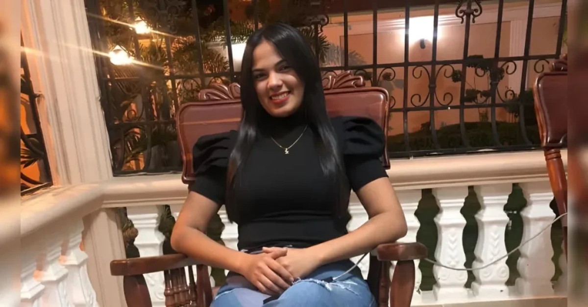 Smiling woman with long dark hair sits in a wooden chair on a balcony at night, wearing a black top and jeans with warm lights behind her.