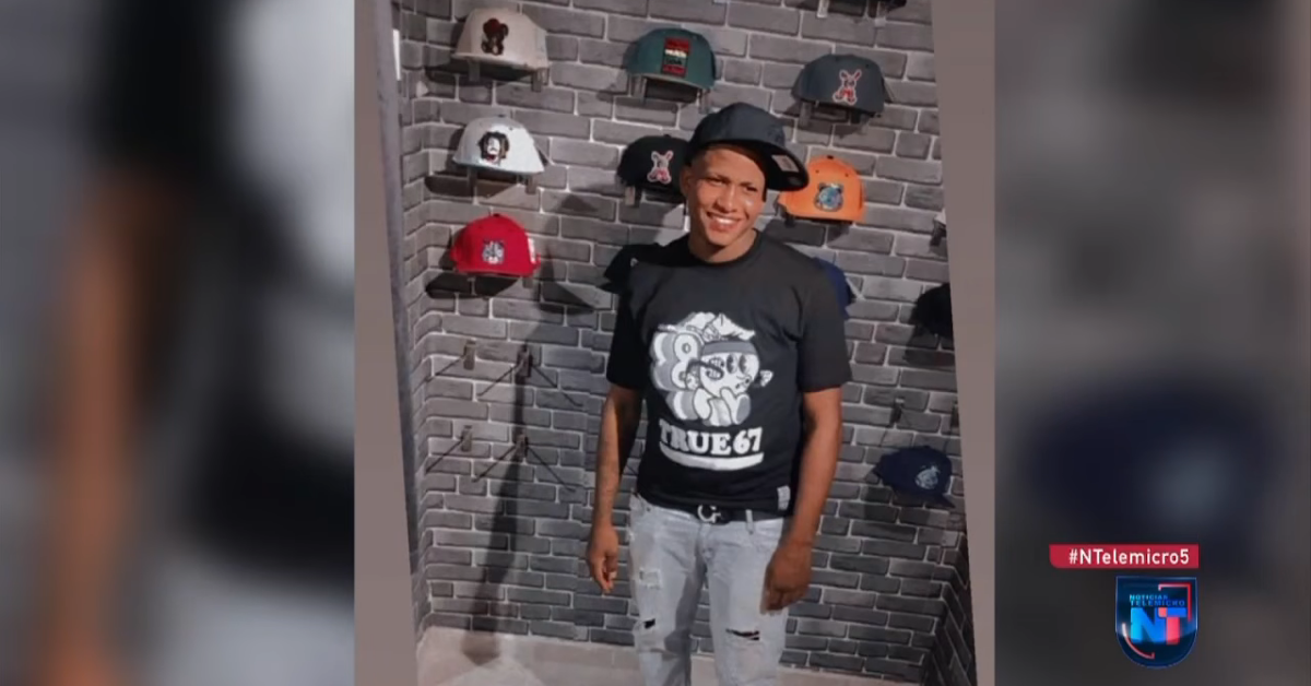 Young man wearing a black graphic T-shirt and light jeans posing in a hat shop with caps on the brick wall behind him.