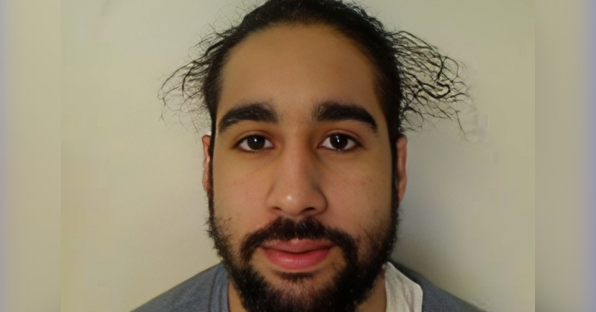 Close-up portrait of a young man with dark hair tied back and a beard, neutral expression against a pale background.
