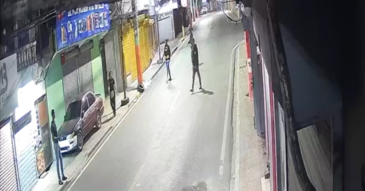 Nighttime city street from a security camera: several people on the sidewalk and road near closed shops and a parked car lining the curb far left.