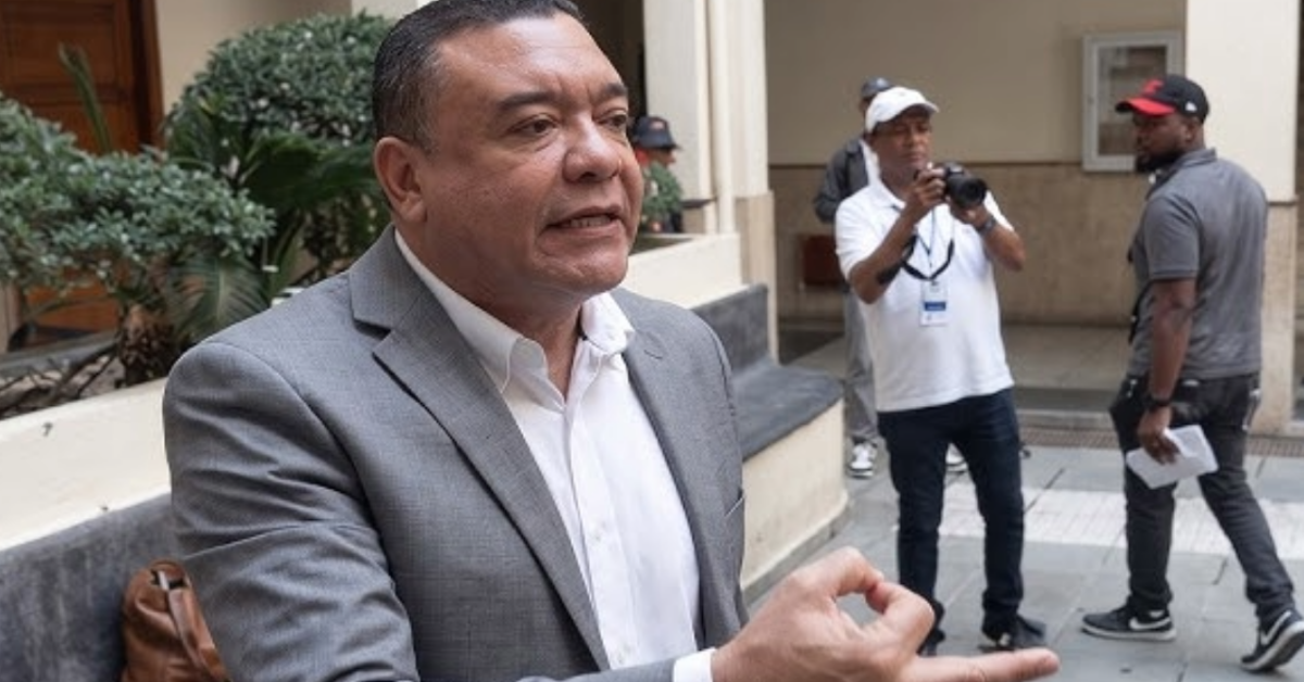 Man in a gray suit speaks outside a building, gesturing with his hands as a small crew films in the background.