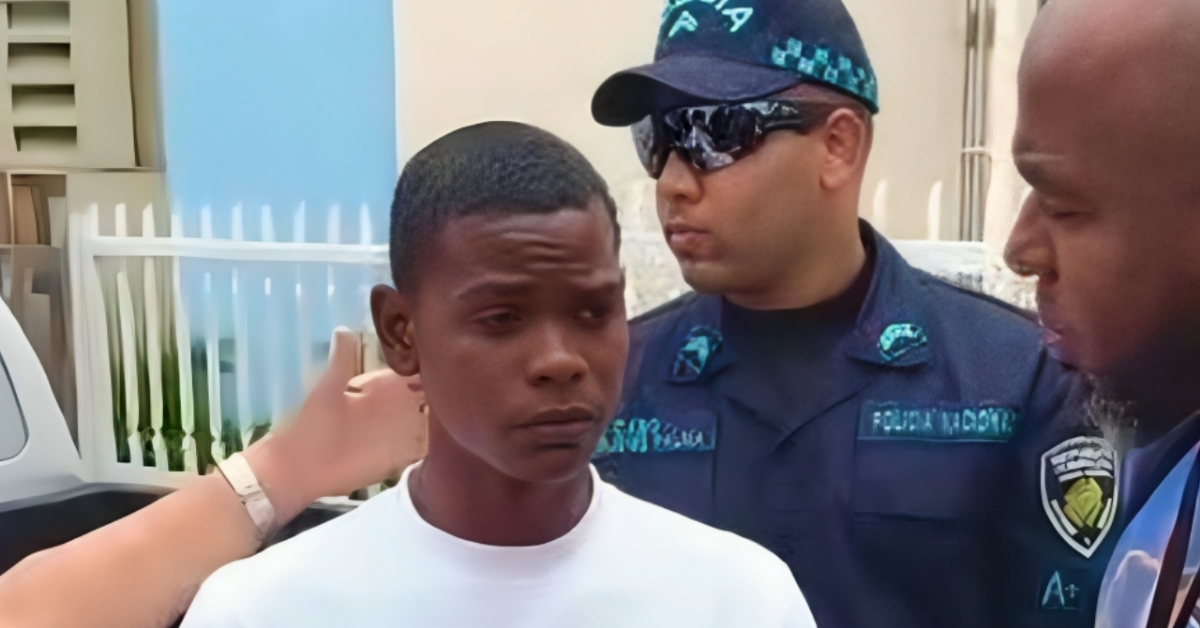 Young man in a white shirt stands with a uniformed security officer wearing sunglasses, outdoors, as another man looks on nearby.