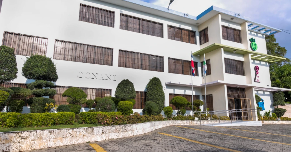 White multi-story building with barred windows, manicured shrubs in front, and flags near the entrance; a paved parking area in the foreground.