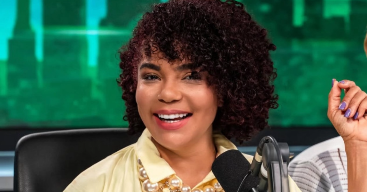 Smiling woman with curly hair in a yellow blouse speaking into a studio microphone in a broadcast setting
