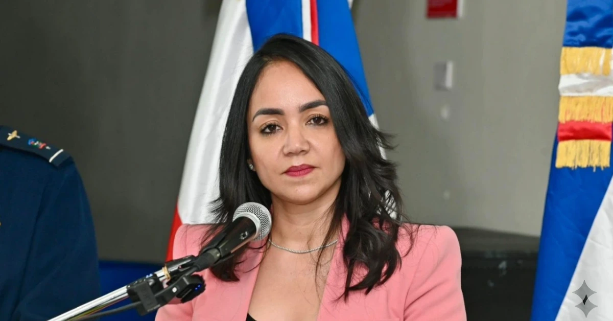Woman in pink blazer speaking into a microphone at a press conference, with flags in the background.