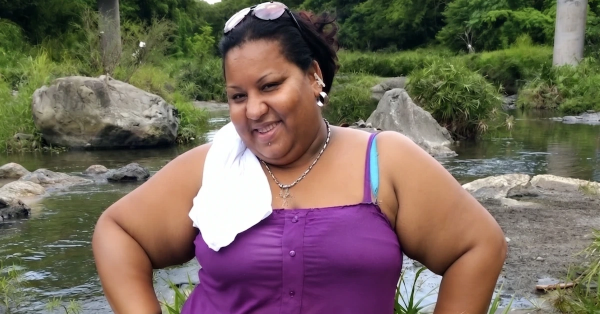Smiling woman in a purple tank top stands by a rocky riverbank with greenery.