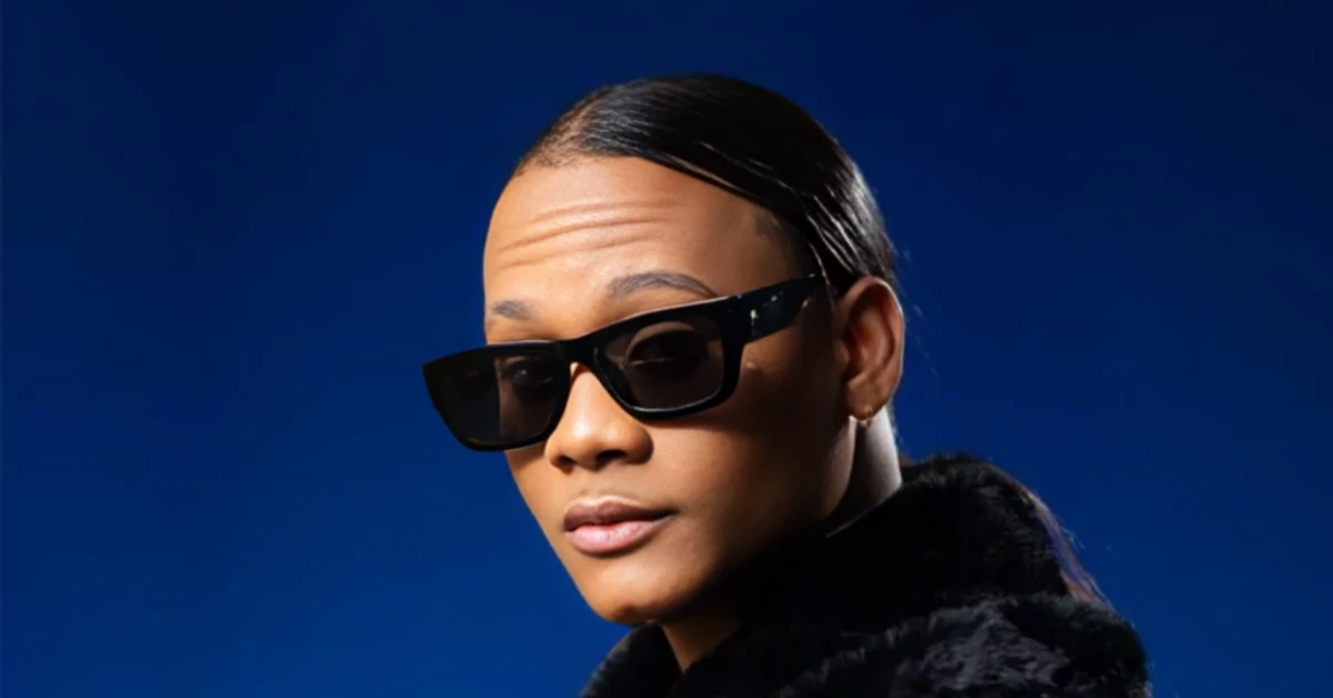 Portrait of a person wearing black sunglasses and a fur-collared coat against a deep blue background.