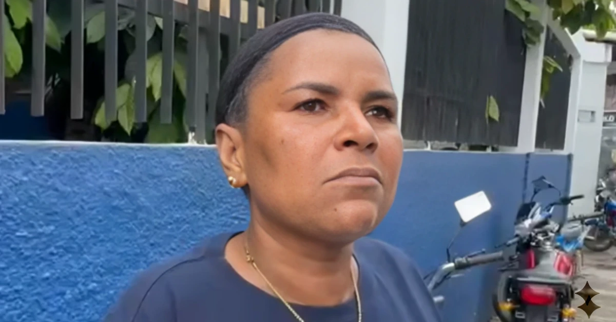 Woman with short hair and a necklace stands beside a blue wall, with a motorcycle visible to the right and greenery in the background.