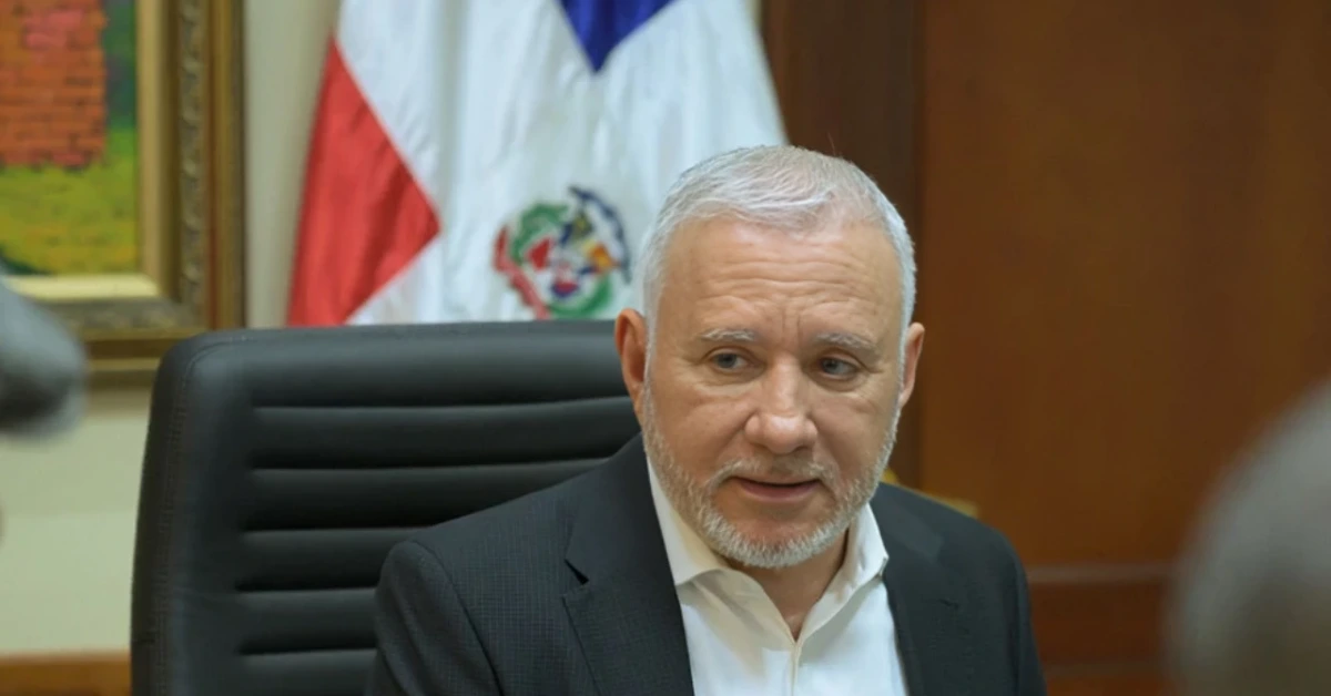Man in a dark suit seated in a meeting room, with a Dominican flag and wooden paneling in the background.