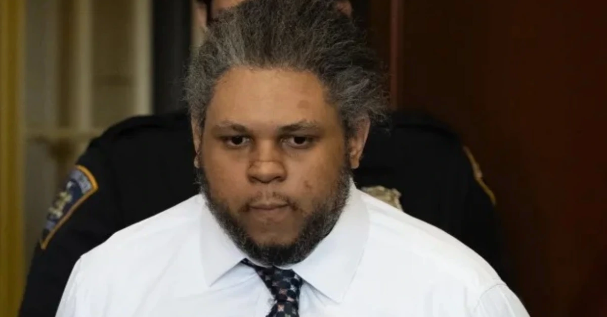Man with gray hair and beard in a white shirt and patterned tie, seated in a courtroom-looking setting with an officer in the background.