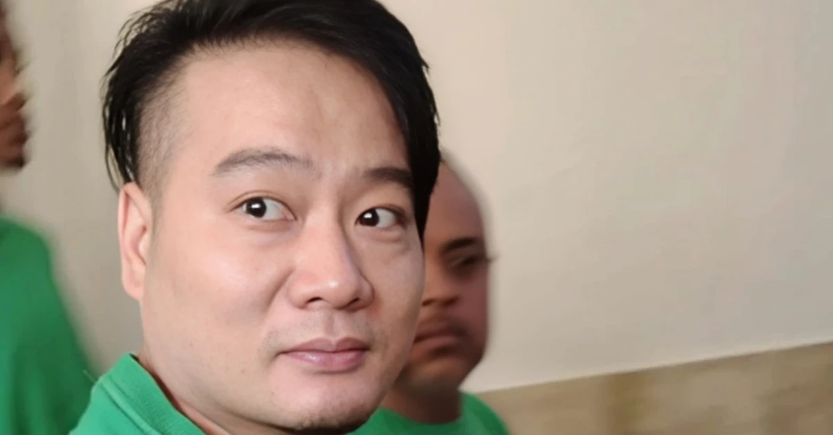 Close-up portrait of a man with short black hair wearing a green shirt, looking toward the camera with a calm expression; a blurred person in green is in the background.