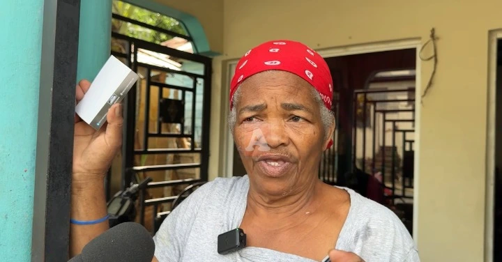 Older woman wearing a red bandana speaks to reporters outside a building, holding a small white box and wearing a clip-on mic.