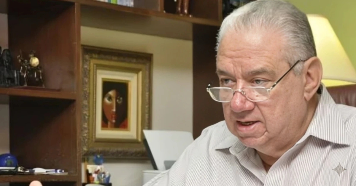 Older man with gray hair and glasses sits at a desk in an office, speaking, with a framed portrait on a bookshelf behind him.