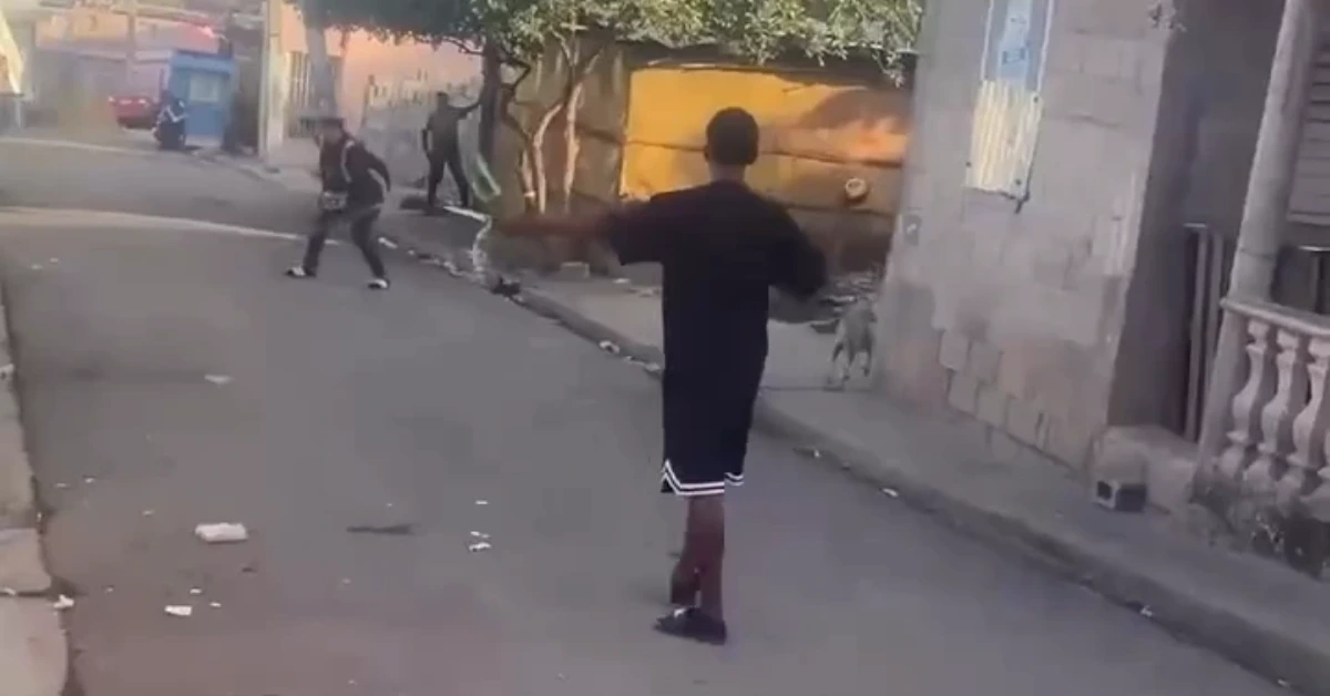Young man in a black shirt and shorts stands in an alley with arms raised as several people fight in the background.