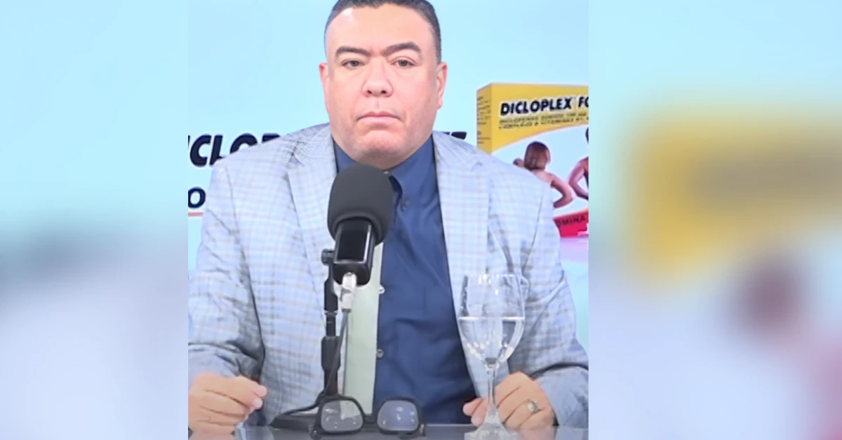 Man in a light checkered blazer and blue shirt sits at a desk, speaking into a wired microphone during a broadcast; a wine glass and sunglasses are on the table, with a product box visible in the background.