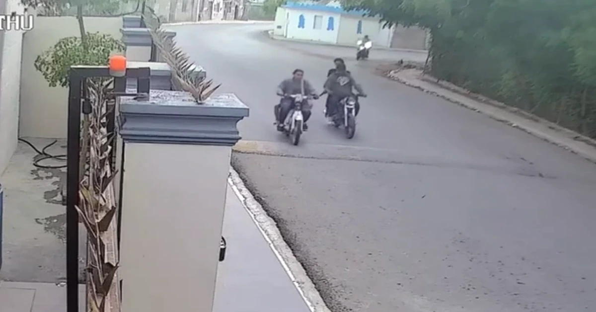 Two motorcyclists ride toward the camera on a quiet residential street, with a gated wall and plants to the left and a blue-roofed building in the distance.