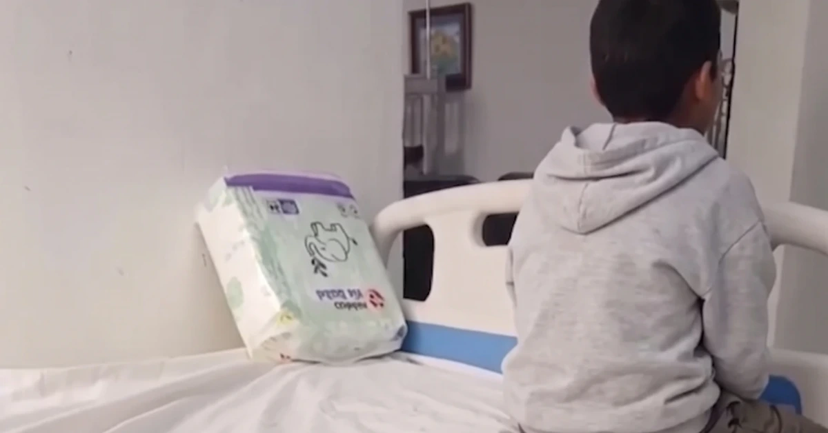 Child in a gray hoodie sits on a hospital bed beside a package of diapers leaning against the wall.