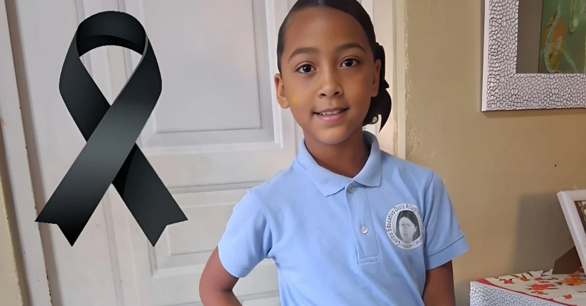 Young girl in a light blue school uniform poses indoors beside a large black awareness ribbon graphic on the wall/doorway.