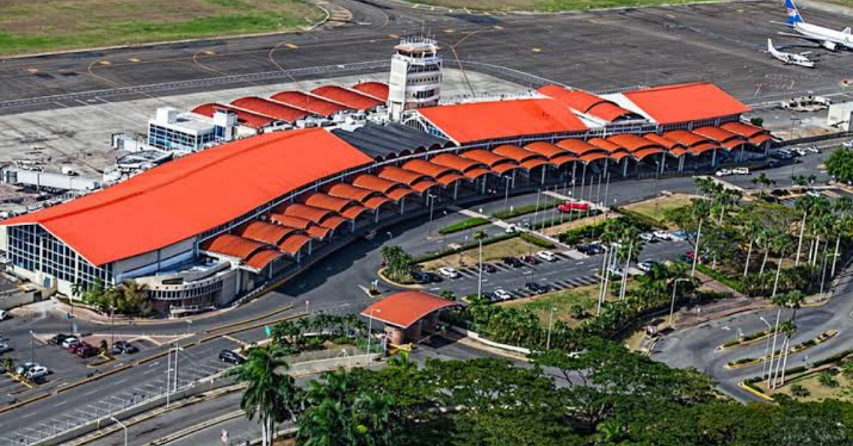 Aerial view of a large airport terminal with a red curved roof and connected concourses (from above). Buildin g features gates and surrounding roads.