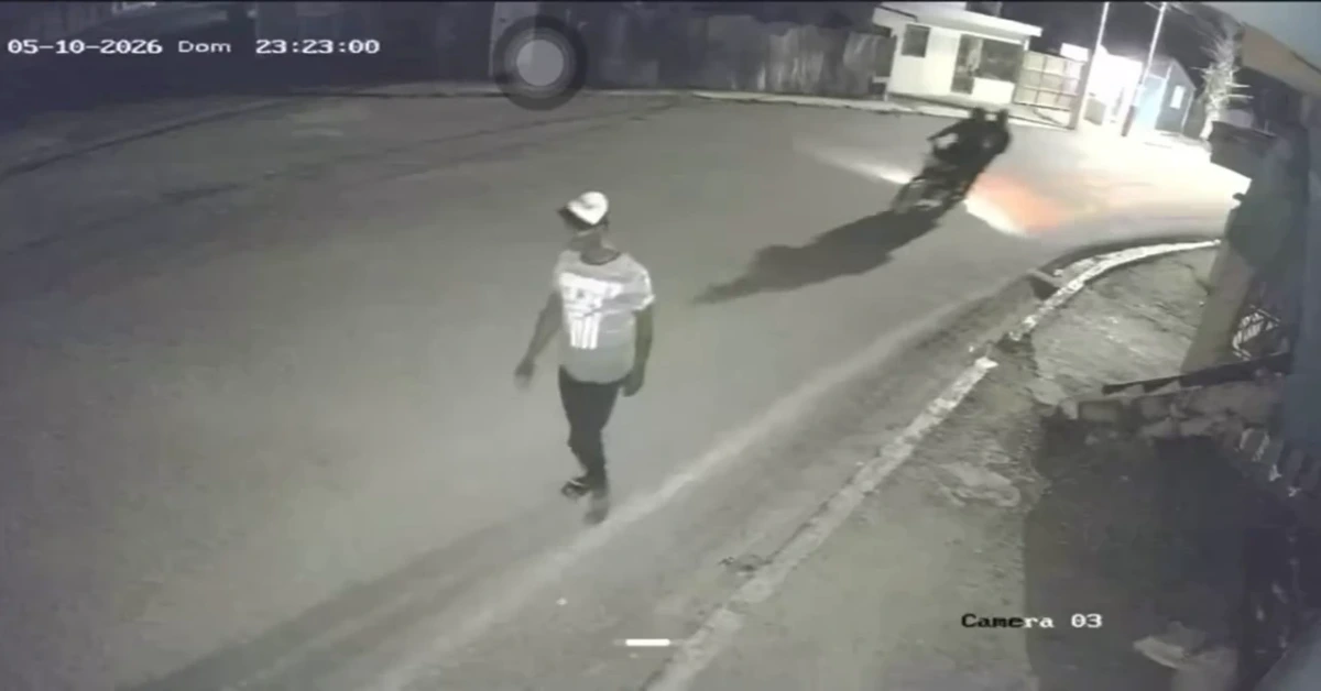 Nighttime security footage of a man in a light hoodie and cap walking on a street, with a motorcycle rider in the distance.