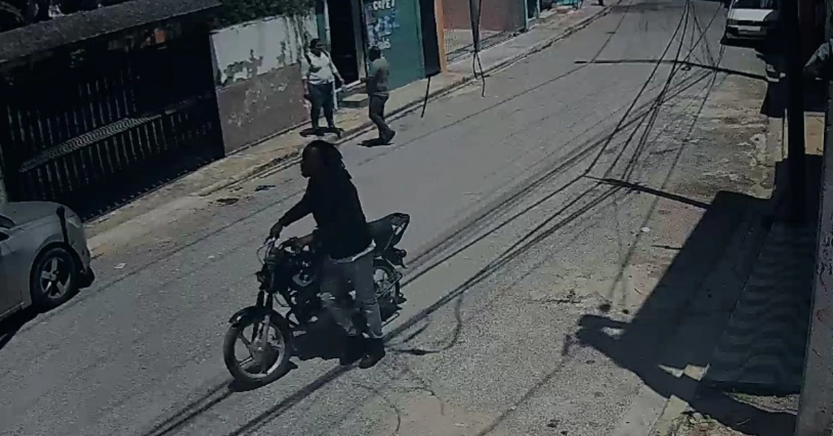 Person on a motorcycle rides down a sunlit urban street; two pedestrians walk on the sidewalk in the background, with utility lines overhead.