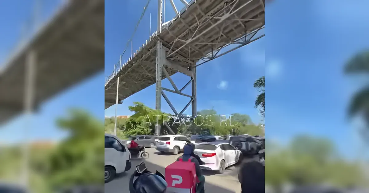 Cars and a motorcycle navigate a congestion under a tall steel bridge on a sunny day.