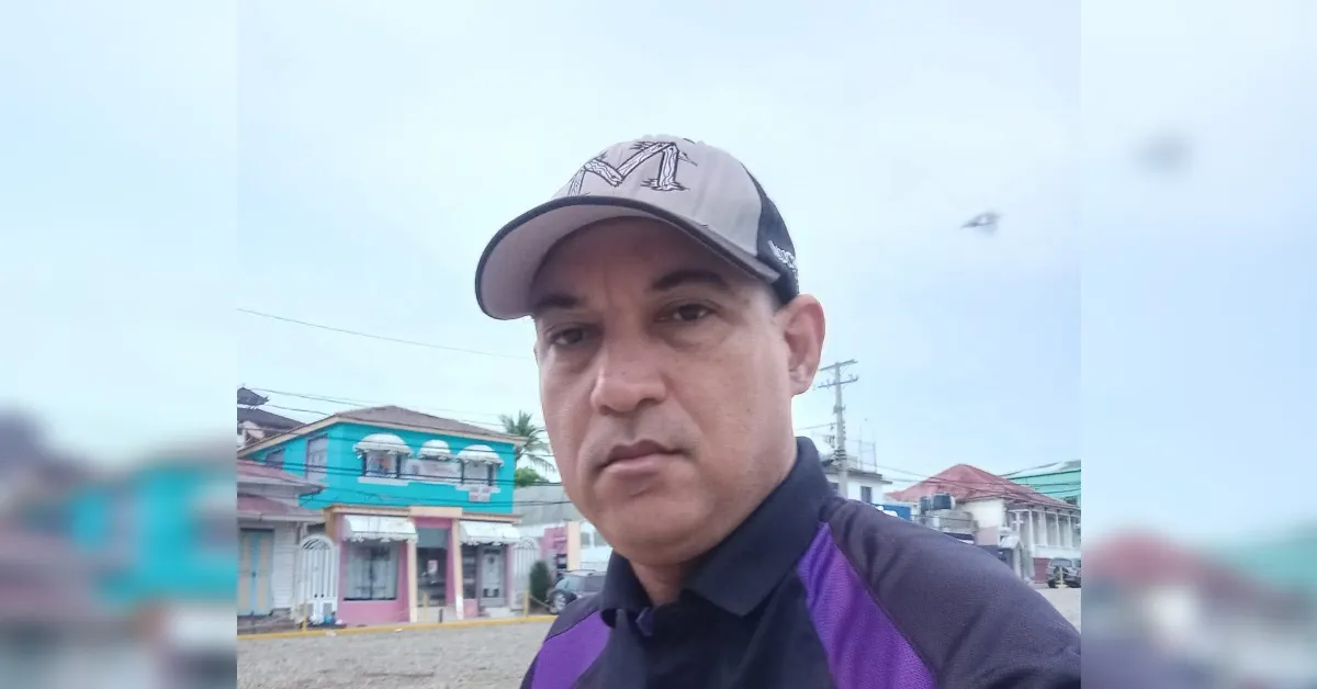 Portrait of a man wearing a light gray cap and purple-black polo shirt, standing on a street with colorful storefronts in the background.