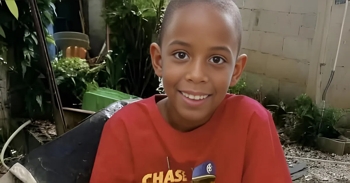 Smiling boy with a shaved head wearing a red T-shirt sits outdoors in a garden or backyard setting.