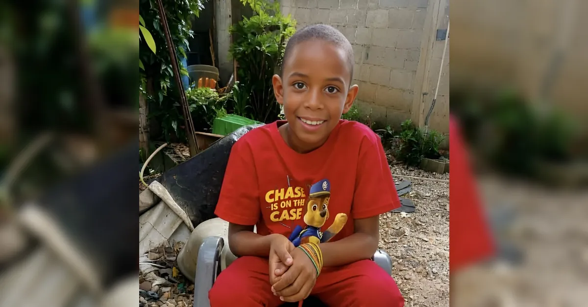 Smiling boy in a red shirt sits on a chair outdoors in a garden setting, hands clasped in his lap, looking at the camera.