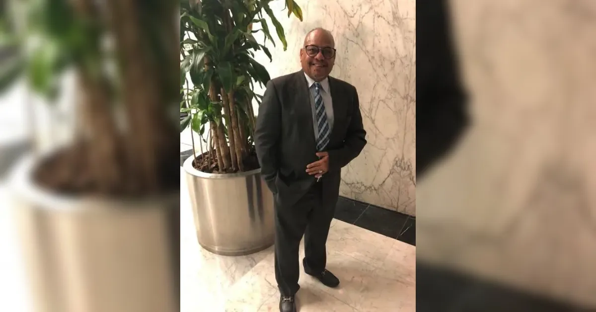 Man in a dark suit and striped tie posing in a marble lobby beside a tall potted plant, smiling at the camera.