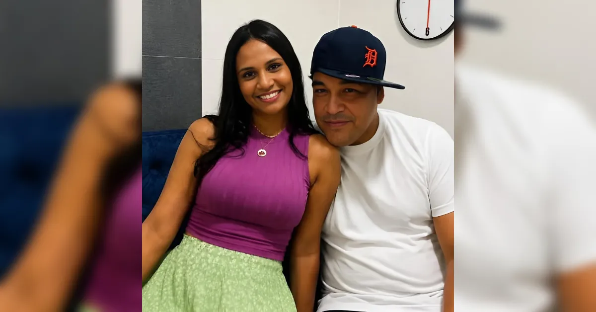 Smiling couple sitting close together indoors: woman in a magenta sleeveless top and light green skirt, man in a white T-shirt and a black Detroit Tigers cap, clock on the wall behind them.