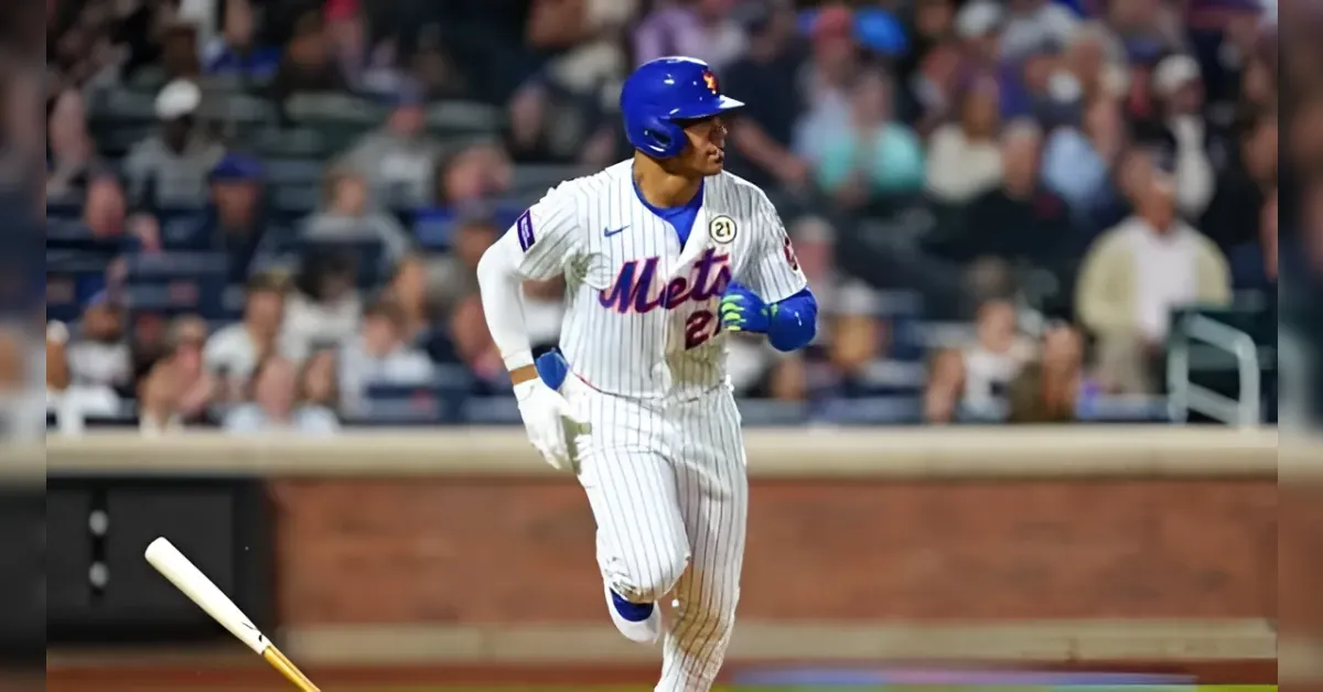 Baseball player in Mets uniform runs on the field as a bat lies on the ground nearby with a blurred stadium crowd in the background.