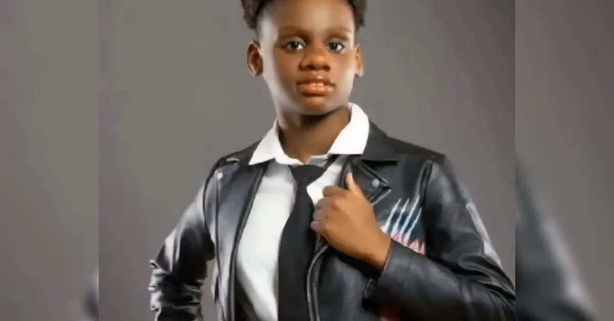 Young person wearing a white collared shirt, black tie, and black leather jacket, posing with one hand on the collar against a gray background.