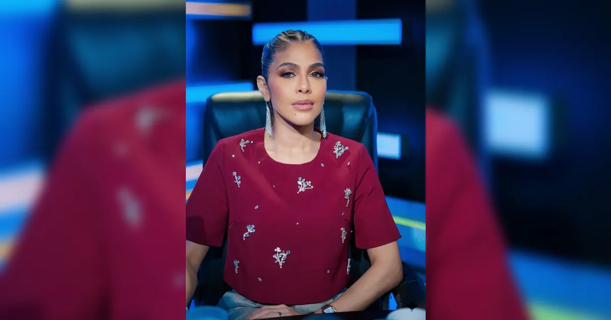 Woman in a red embellished top sits in a studio chair with blue stage lights in the background, looking at the camera.