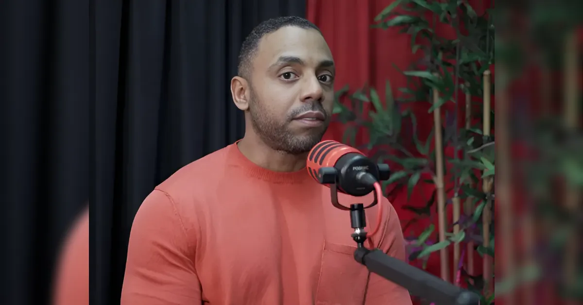 Man in an orange shirt speaks into a red microphone on a stand during a podcast recording set with a dark curtain behind and plants nearby.