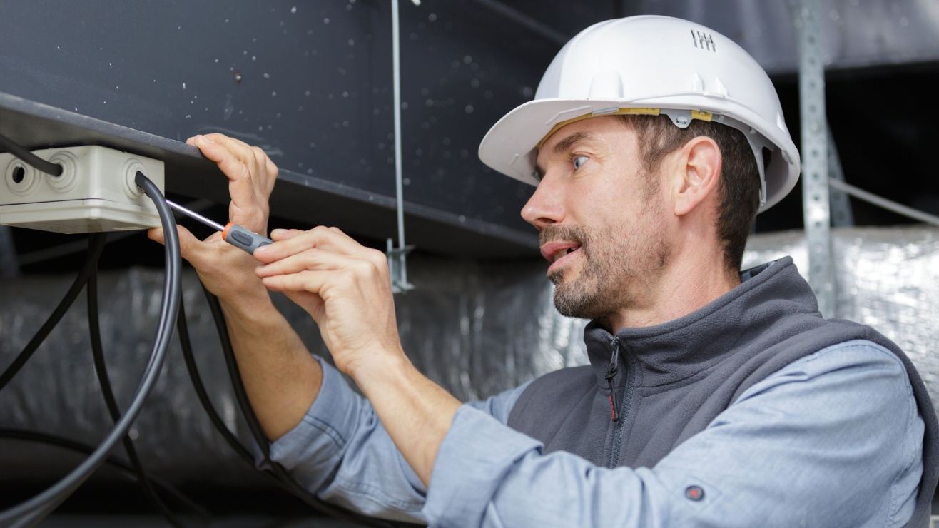 Construction worker wearing a white hard hat uses a screwdriver to adjust wiring under a metal structure, outdoors/industrial setting