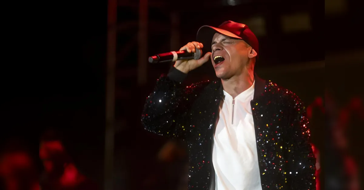 Male singer wearing a red cap and a black sequined jacket, singing into a microphone on stage at a concert.