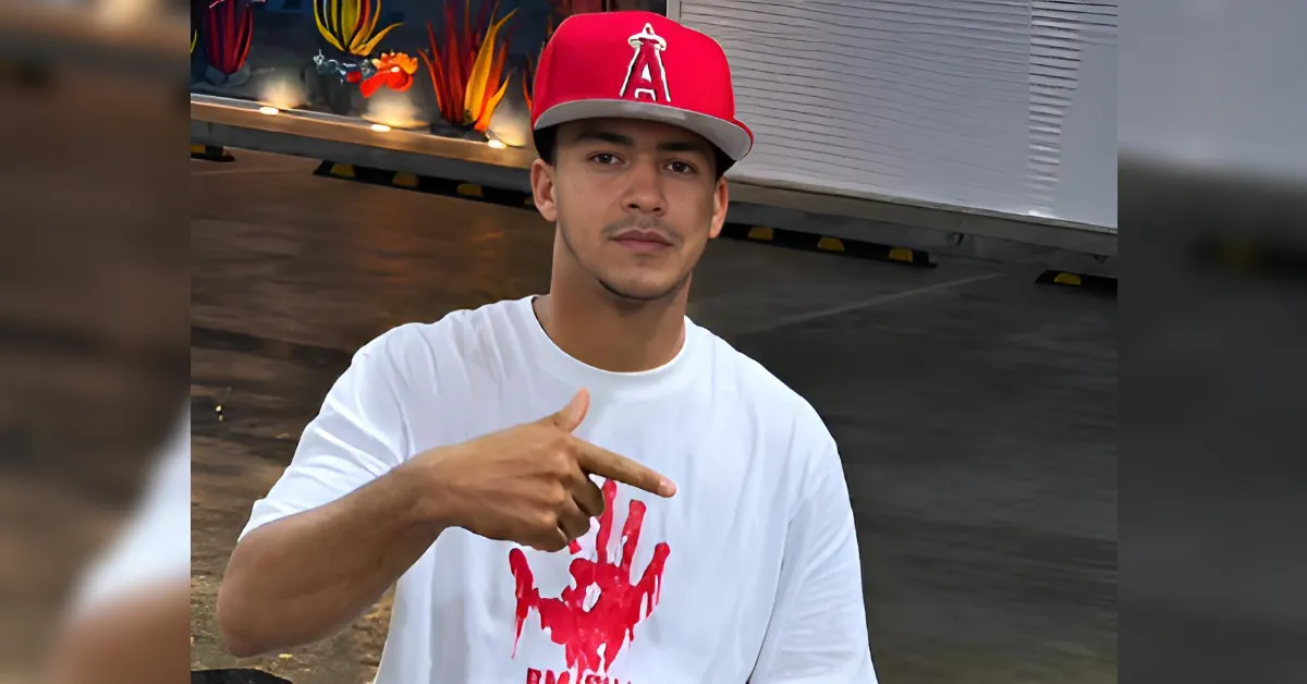 Man in a white T-shirt with a red handprint graphic, wearing a red cap, posing outdoors and pointing sideways with his right hand, urban backdrop behind him.