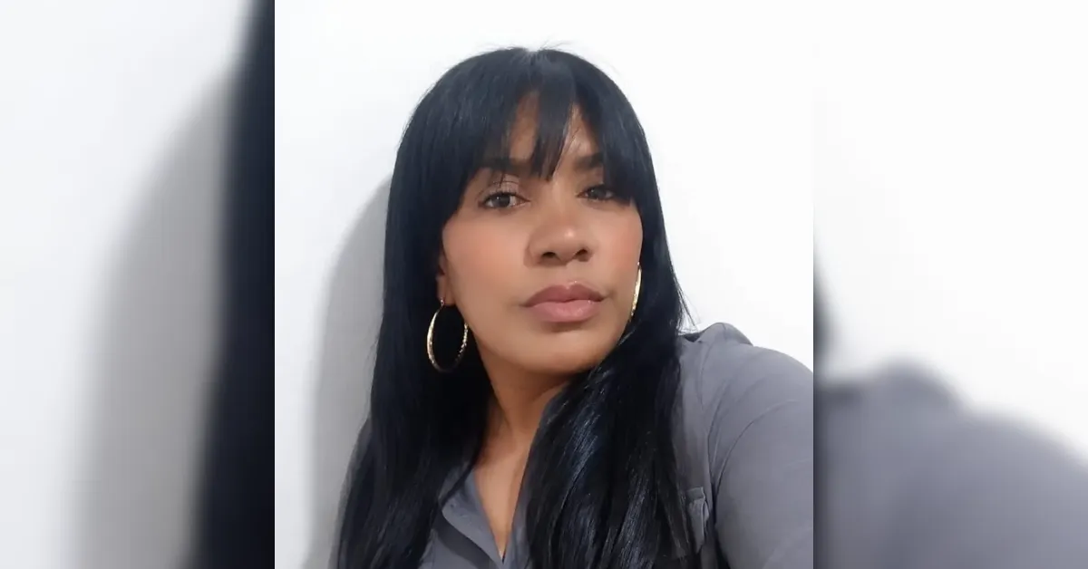 Woman with long black hair and bangs, gold hoop earrings, wearing a gray top, posing against a white wall and looking at the camera.