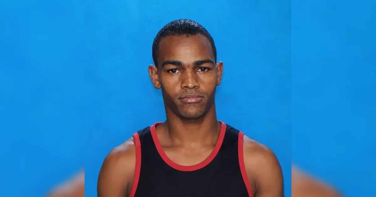 Portrait of a young man with short hair, wearing a black tank top with red trim, posed against a solid blue background with a neutral expression.