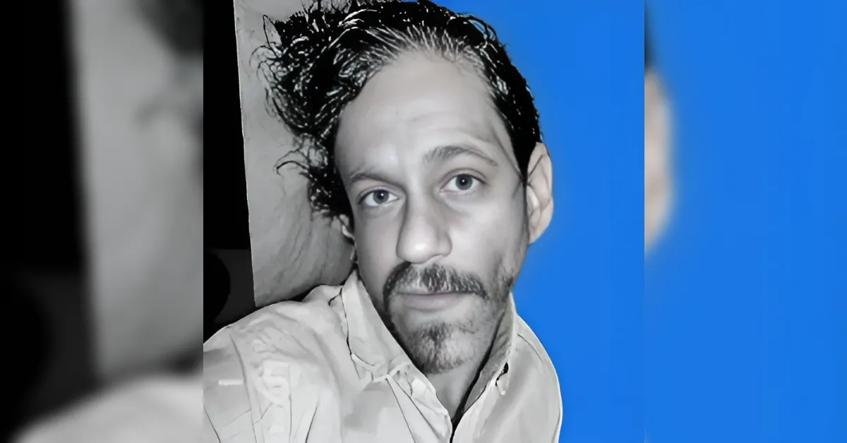 Close-up portrait of a man with damp hair and stubble, facing the camera against a blue background.